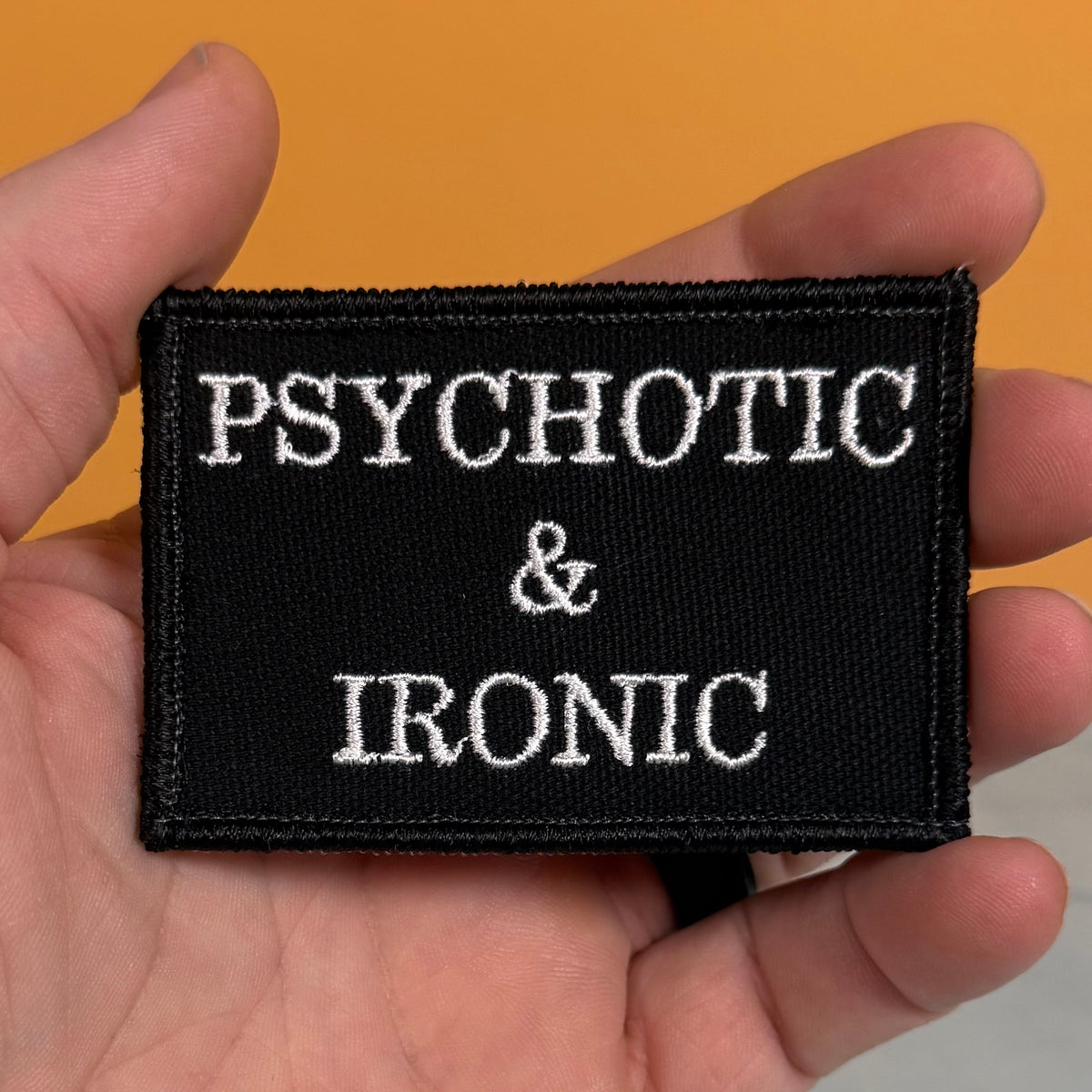 A black rectangular embroidered patch with the words 'PSYCHOTIC & IRONIC' in white lettering, held in a person's hand against a solid orange background.