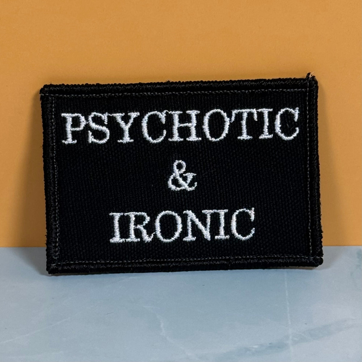 A rectangular black embroidered patch with the words 'PSYCHOTIC & IRONIC' in white capital letters, photographed against a warm orange background and a pale blue-gray surface.