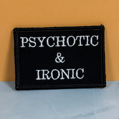 A rectangular black embroidered patch with the words 'PSYCHOTIC & IRONIC' in white capital letters, photographed against a warm orange background and a pale blue-gray surface.