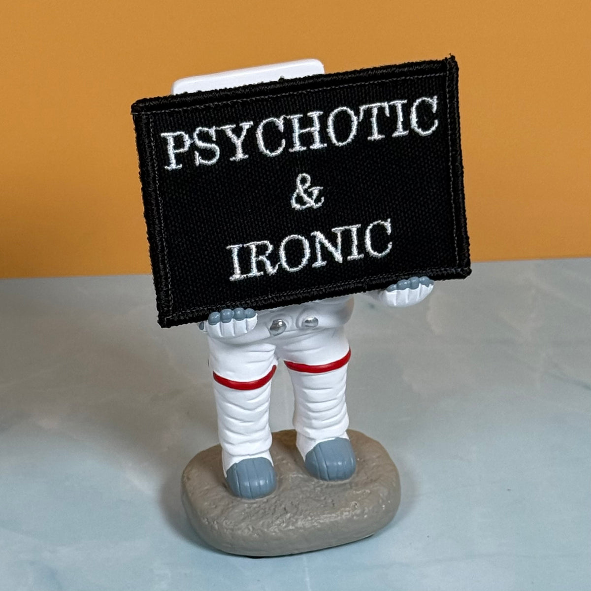 A novelty astronaut figurine holding a black rectangular patch with white embroidered text reading 'PSYCHOTIC & IRONIC', set against a two-tone orange and gray background.