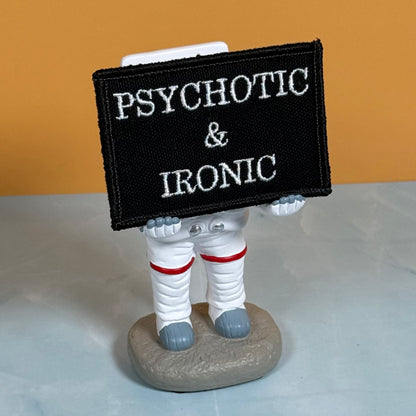 A novelty astronaut figurine holding a black rectangular patch with white embroidered text reading 'PSYCHOTIC & IRONIC', set against a two-tone orange and gray background.
