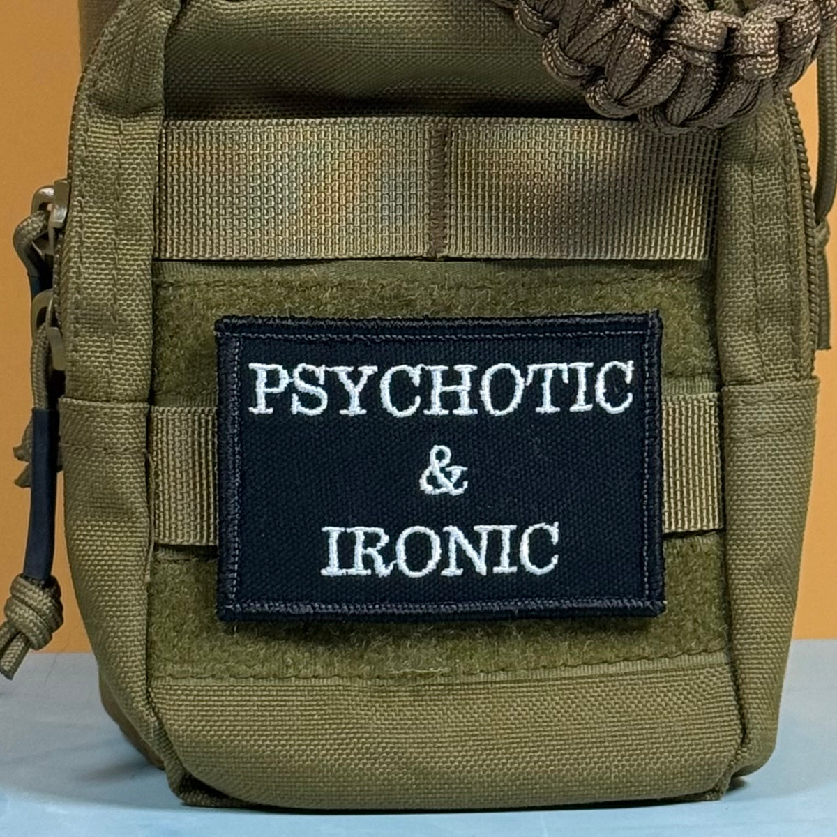 An olive drab tactical pouch with a black embroidered patch that reads 'PSYCHOTIC & IRONIC' in white text, shown against a solid orange background.