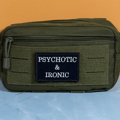 An olive green tactical pouch with a black rectangular patch that reads 'PSYCHOTIC & IRONIC' in white text, set against a solid orange background and light blue surface.