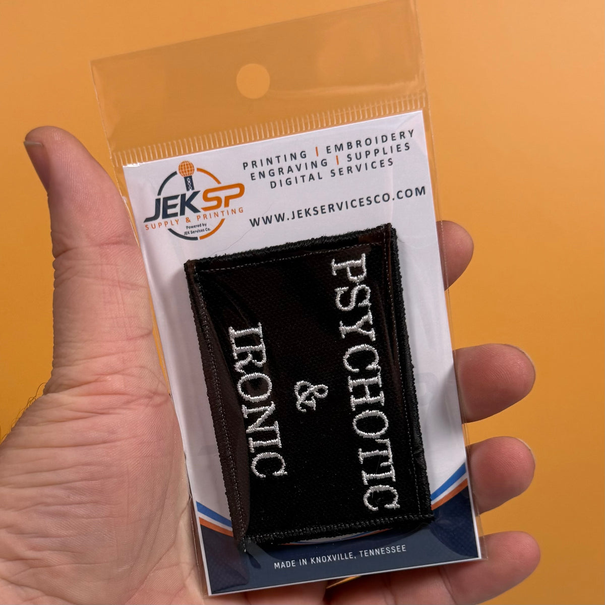 A hand holds a small embroidered patch in clear retail packaging against a solid orange background. The patch reads 'PSYCHOTIC & IRONIC' in white on black, and the package card shows the brand Jeksp Supply Services.