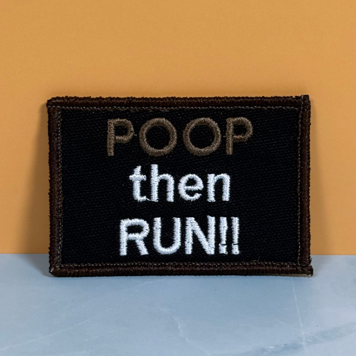 A rectangular embroidered patch with the text 'POOP then RUN!!' on a black background, placed against a warm orange wall and a light blue-gray surface.