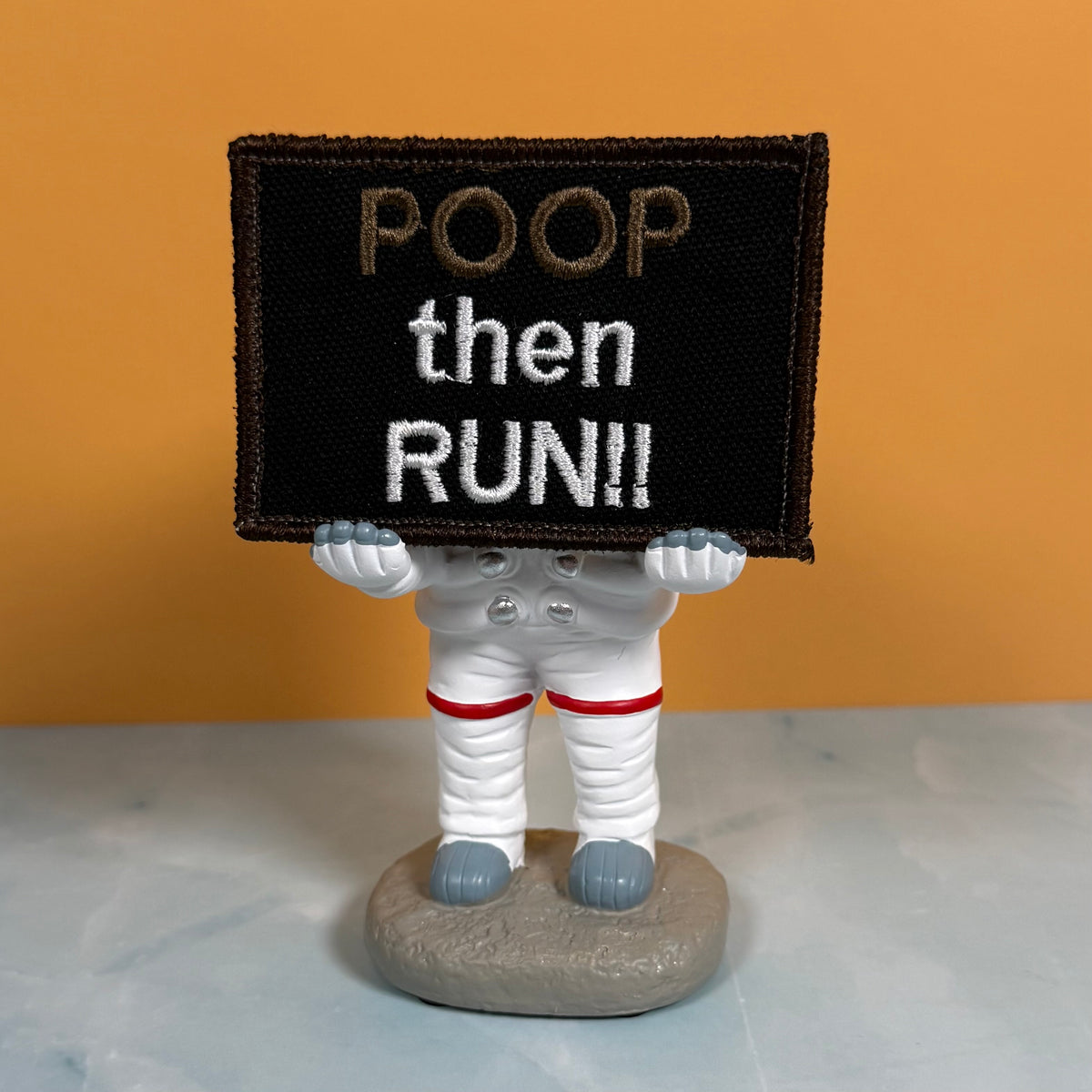 An astronaut figurine holding a black rectangular sign that reads 'POOP then RUN!!' in embroidered text, set against an orange background and light gray surface.
