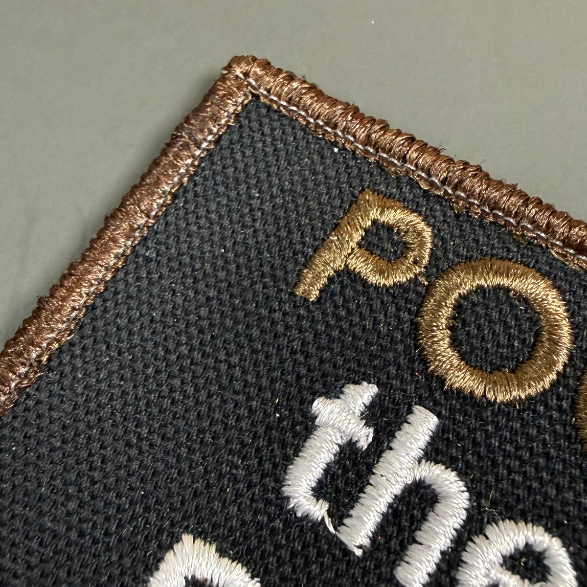Close-up of a black embroidered patch with a brown stitched border, gold lettering, and white lettering on a neutral gray background.