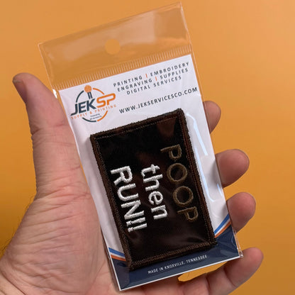 A hand holds a packaged embroidered patch that reads 'POOP then RUN!!' in white and gold on a dark brown background. The patch is sealed in a clear plastic sleeve with a branded JEKSP backing card, against a solid orange background.