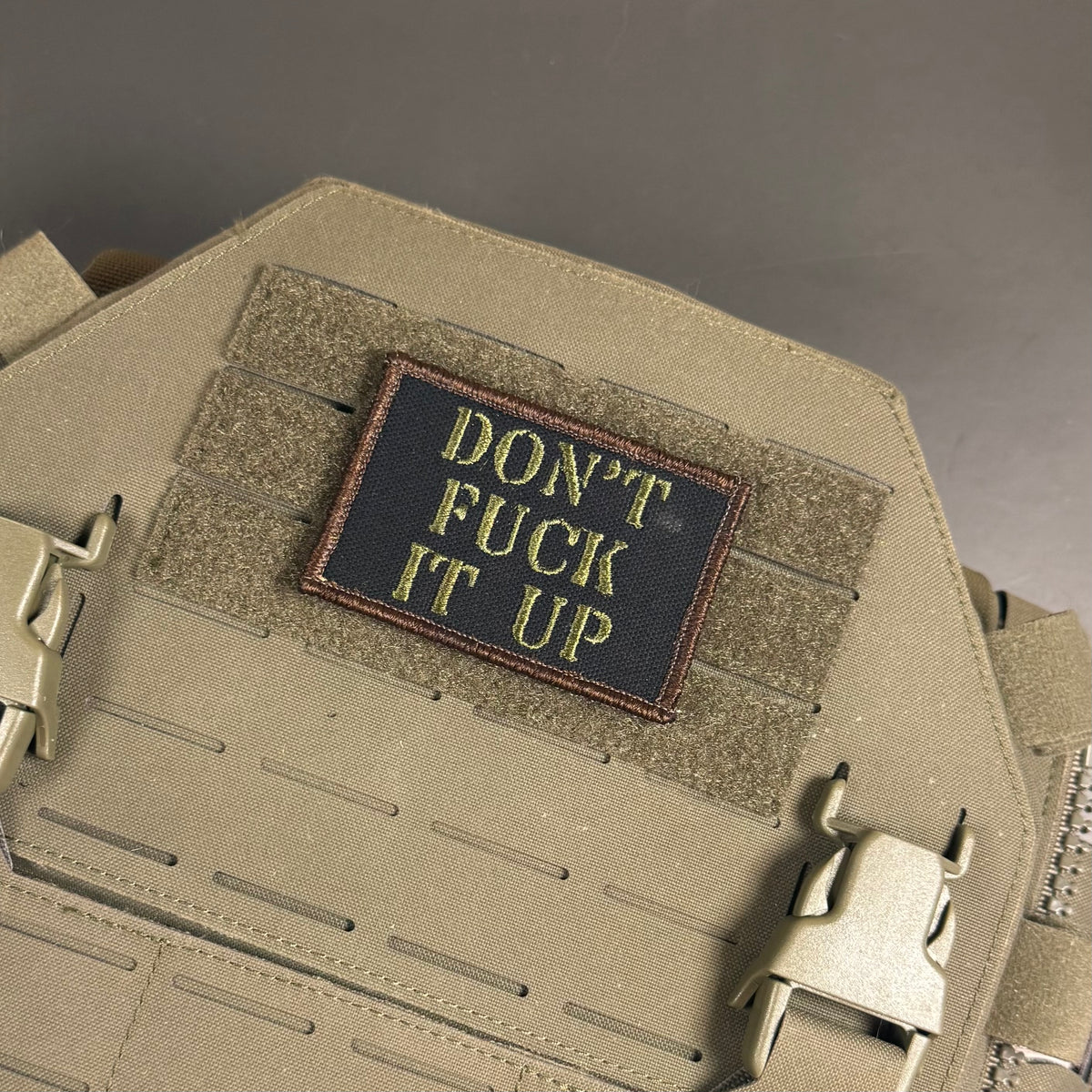 A rectangular embroidered patch with the text 'DON'T FUCK IT UP' in yellow on a black background, sewn onto a tan tactical vest with Velcro and quick-release buckles.