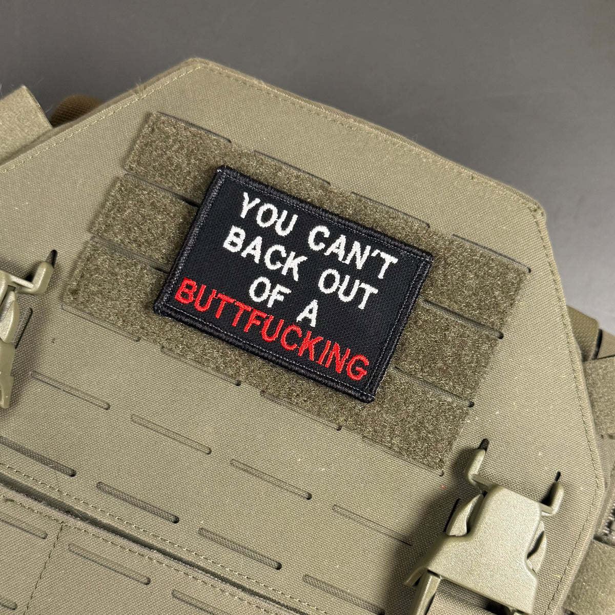 A rectangular embroidered patch with the text 'YOU CAN'T BACK OUT OF A BUTTFUCKING' in white and red on a black background, attached to tan tactical webbing on a beige vest or plate carrier.