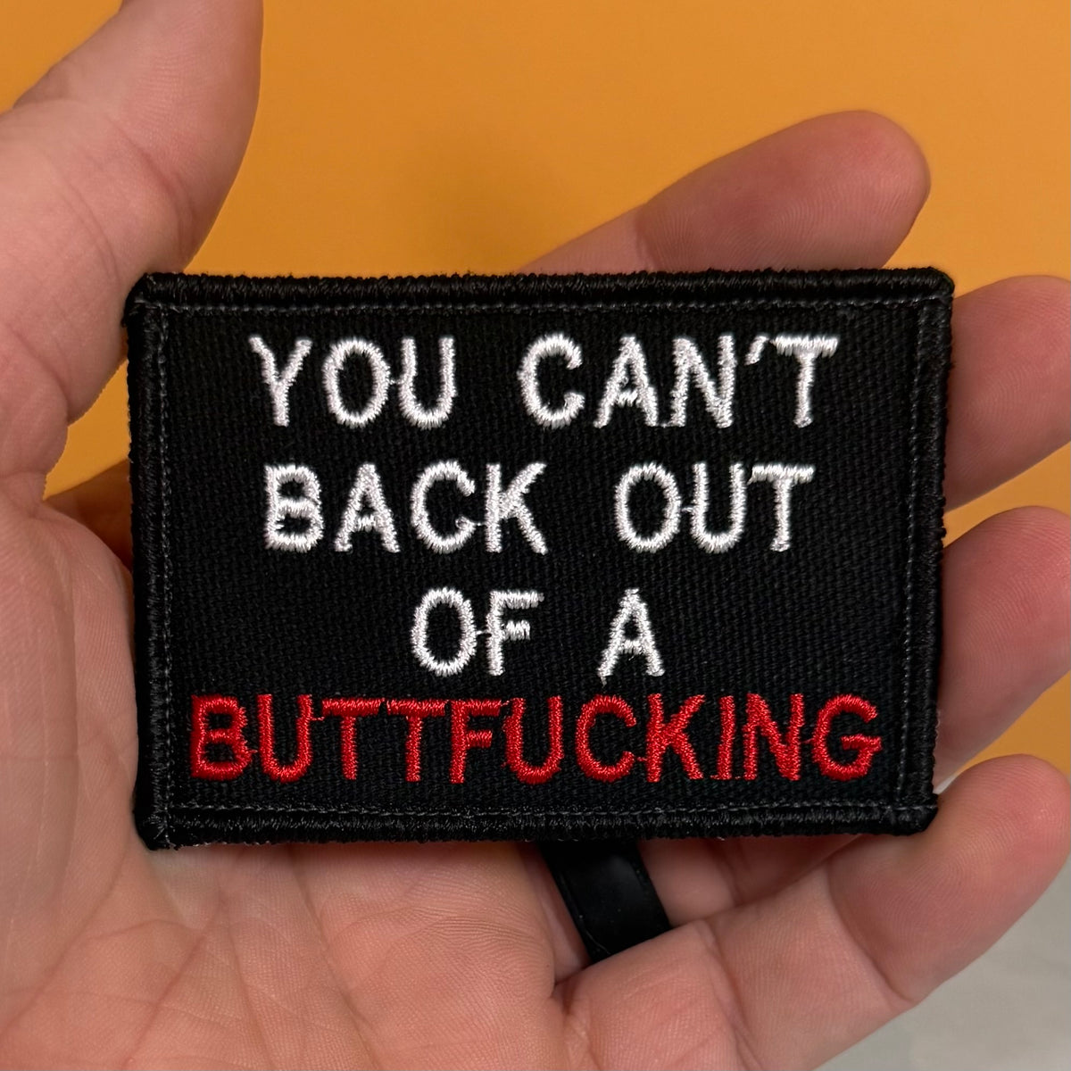 A hand holding a rectangular black embroidered patch with the text 'YOU CAN'T BACK OUT OF A BUTTFUCKING' in white and red lettering, against a solid orange background.