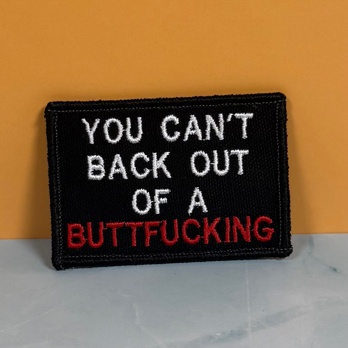 A rectangular black embroidered patch with the text 'YOU CAN'T BACK OUT OF A BUTTFUCKING' in white and red lettering, photographed against a mustard yellow background and light gray surface.