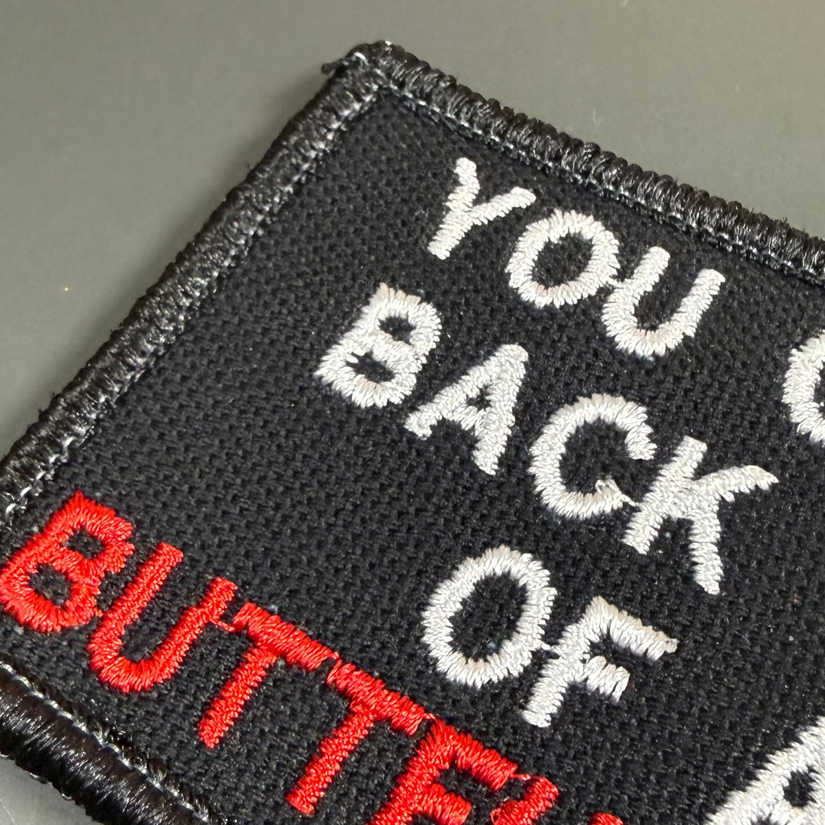 Close-up of a black embroidered patch with white and red text reading 'YOU BACK OF BUTTERFLY' on a dark gray background.