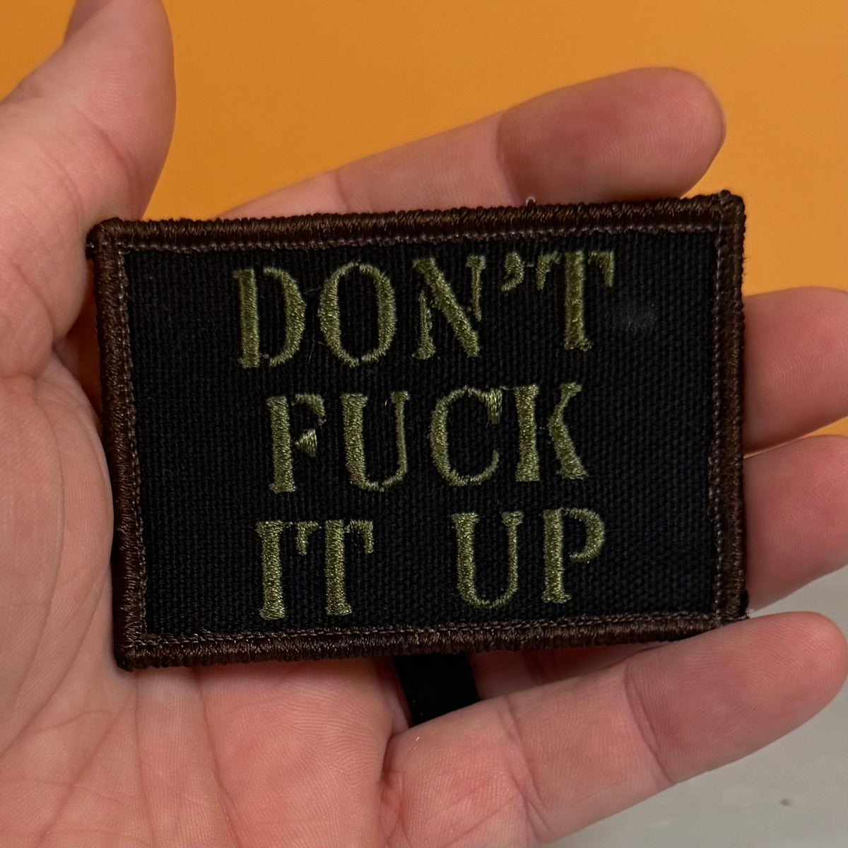 A rectangular embroidered patch held in a hand, featuring the text 'DON'T FUCK IT UP' in olive green lettering on a black background with a dark brown stitched border.