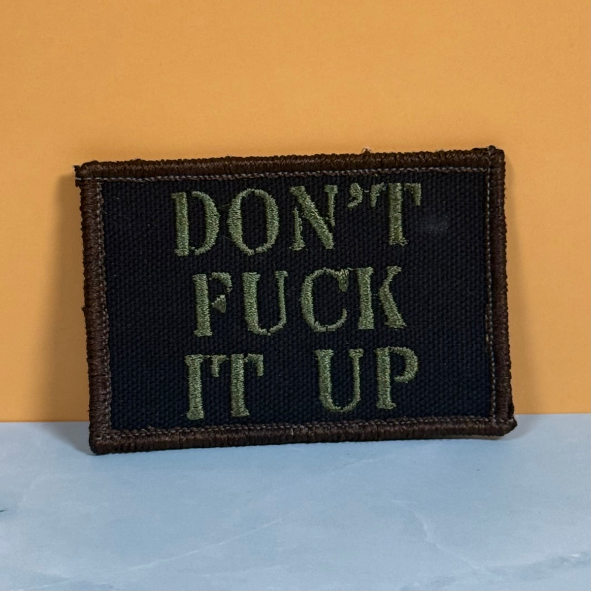 A rectangular embroidered patch with the text 'DON'T FUCK IT UP' in olive green on a black background, finished with a brown stitched border, leaning against a yellow wall on a light blue surface.