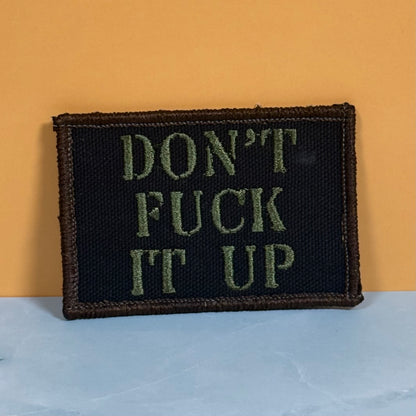 A rectangular embroidered patch with the text 'DON'T FUCK IT UP' in olive green on a black background, finished with a brown stitched border, leaning against a yellow wall on a light blue surface.