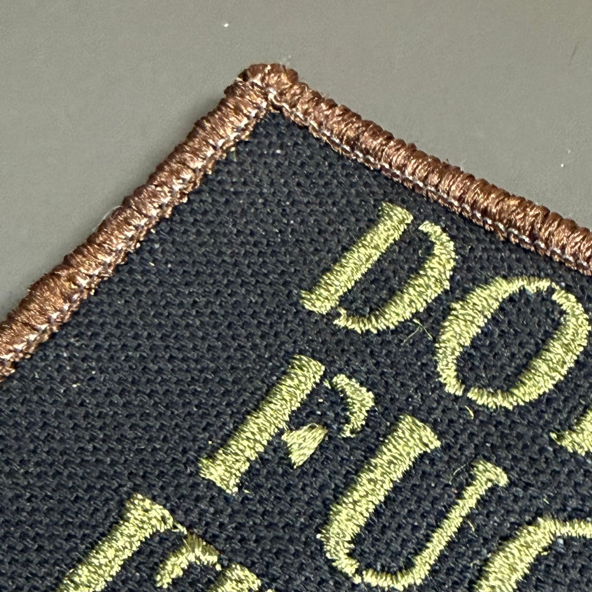 Close-up of a black embroidered patch with a brown stitched border and light olive green lettering on a gray background.