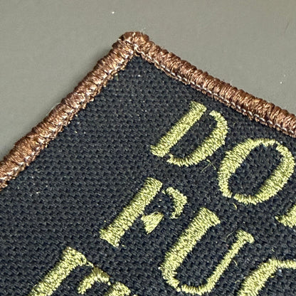 Close-up of a black embroidered patch with a brown stitched border and light olive green lettering on a gray background.