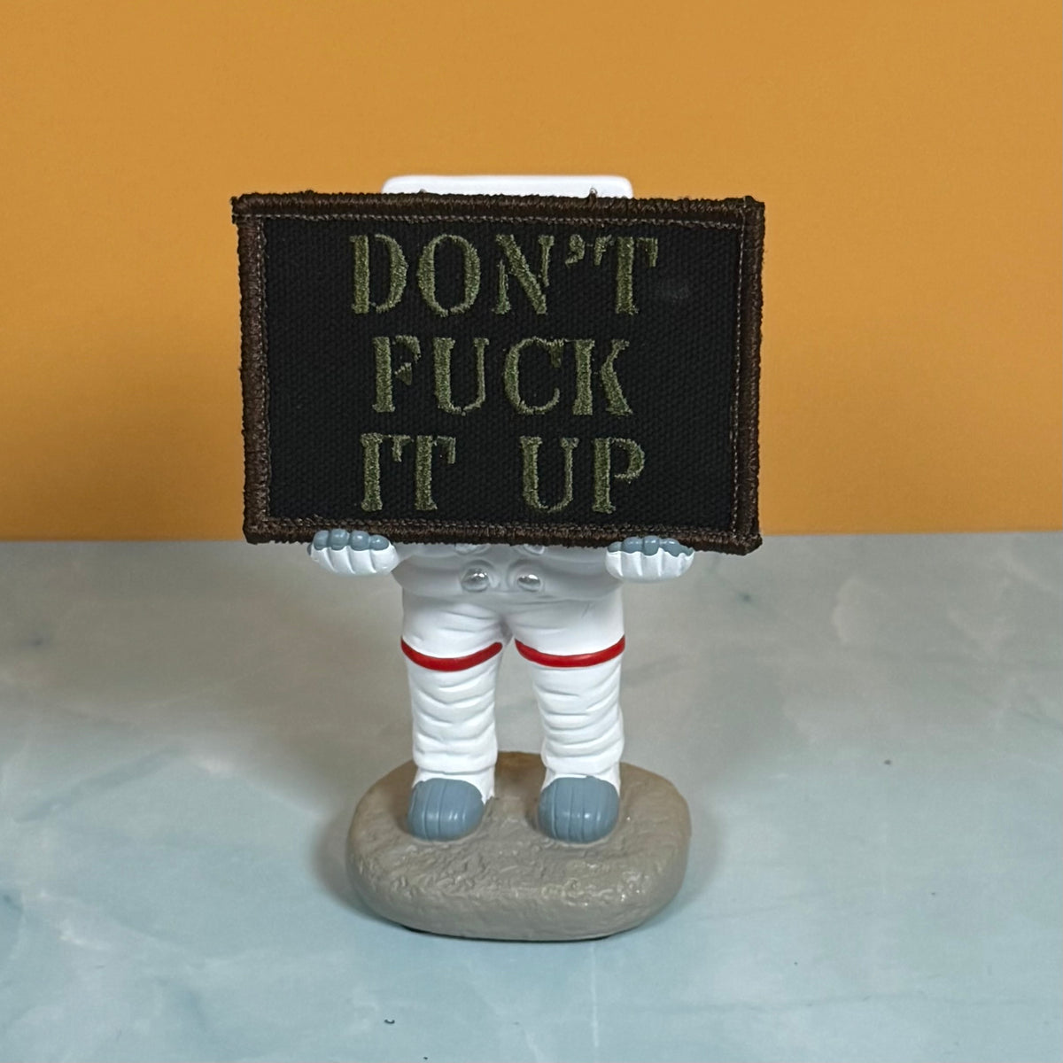 An embroidered patch with the text 'DON'T FUCK IT UP' in olive green letters on a black background, held by a small astronaut figurine on a gray base against a two-tone orange and light gray background.