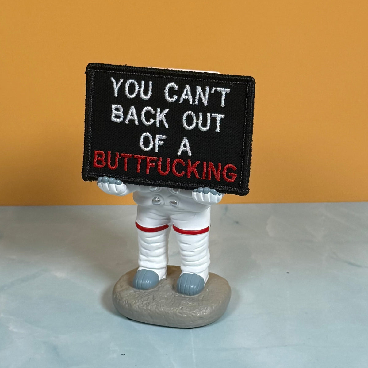 A novelty astronaut figurine holding a black rectangular sign with the embroidered text 'YOU CAN'T BACK OUT OF A BUTTFUCKING'. The figurine is dressed in a white spacesuit with red stripes and gray boots, standing on a gray rock base against a solid orange background and light gray surface.
