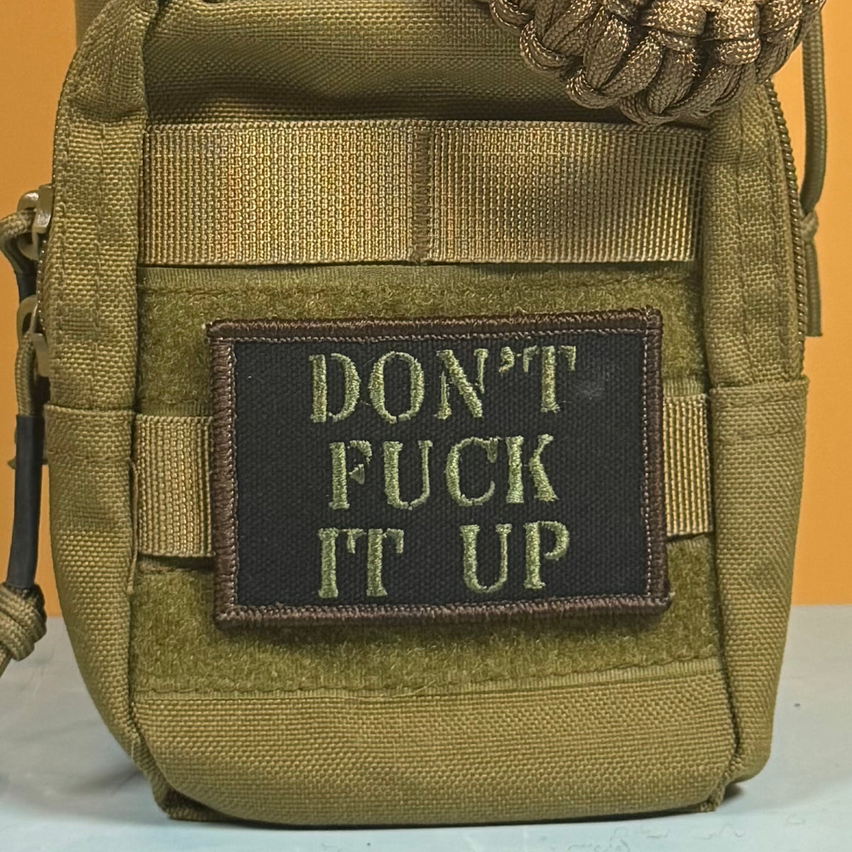 An olive drab tactical pouch with a black rectangular patch that reads 'DON'T FUCK IT UP' in olive drab embroidery, shown against a solid orange background.