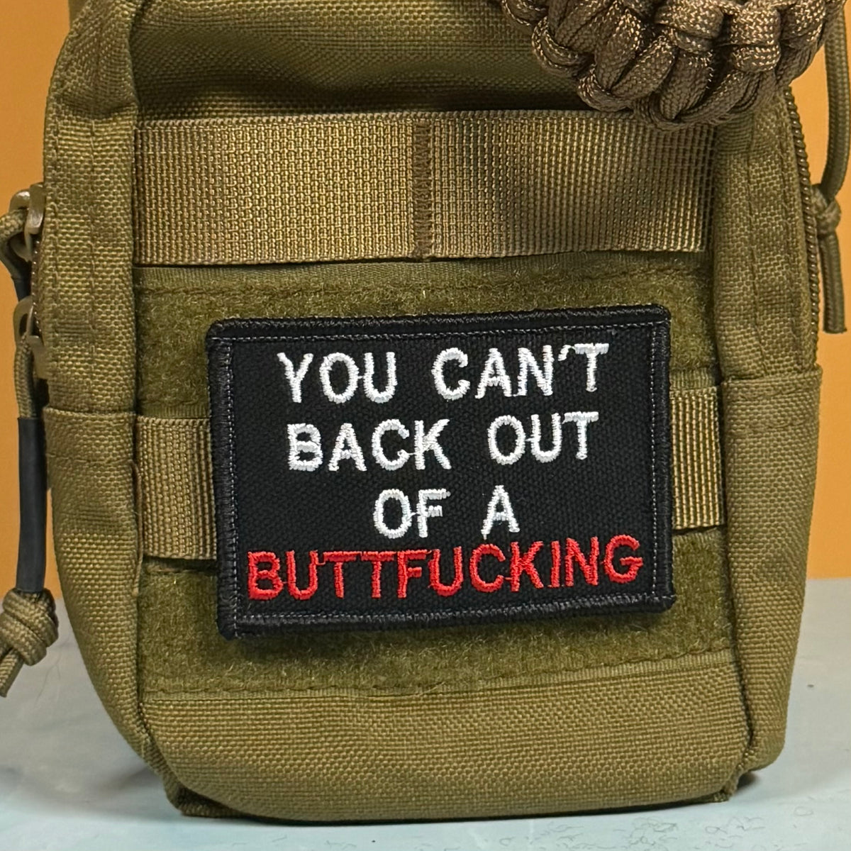 An olive green tactical pouch featuring a black rectangular embroidered patch with the text 'YOU CAN'T BACK OUT OF A BUTTFUCKING' in white and red lettering, set against a solid orange background.