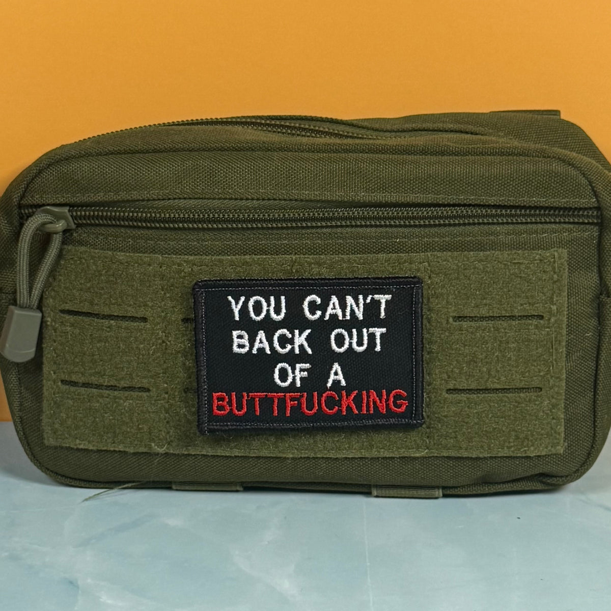 An olive green tactical pouch with a black embroidered patch featuring the text 'YOU CAN'T BACK OUT OF A BUTTFUCKING' in white and red lettering, set against a solid orange background.
