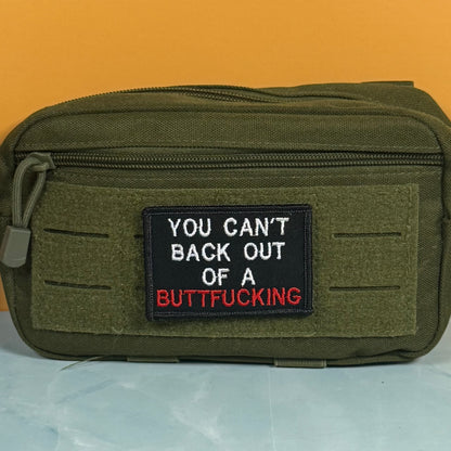 An olive green tactical pouch with a black embroidered patch featuring the text 'YOU CAN'T BACK OUT OF A BUTTFUCKING' in white and red lettering, set against a solid orange background.