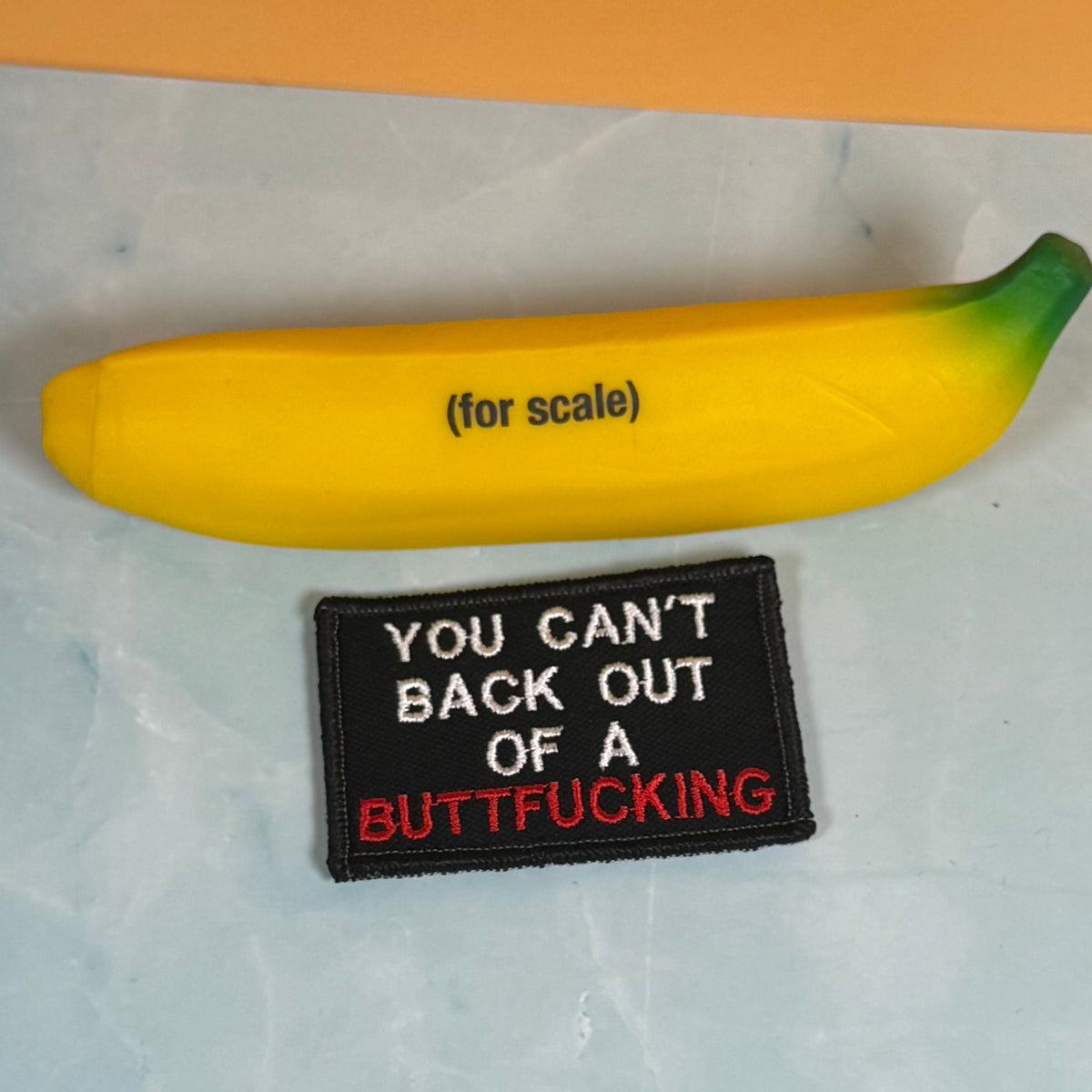 A novelty yellow banana-shaped object with green tips and the text '(for scale)' above a black embroidered patch that reads 'YOU CAN'T BACK OUT OF A BUTTFUCKING'.