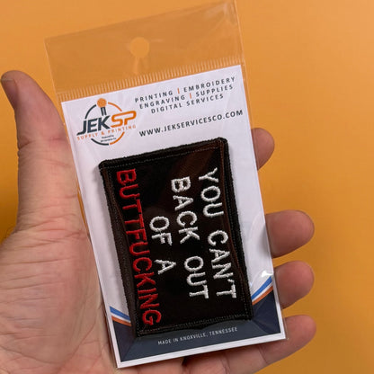 A hand holding a packaged embroidered patch that reads 'BUTTFUCKING' in red and white, with 'YOU CAN'T BACK OUT OF A' in white. The patch is in clear plastic packaging with a branded card header, against a solid orange background.
