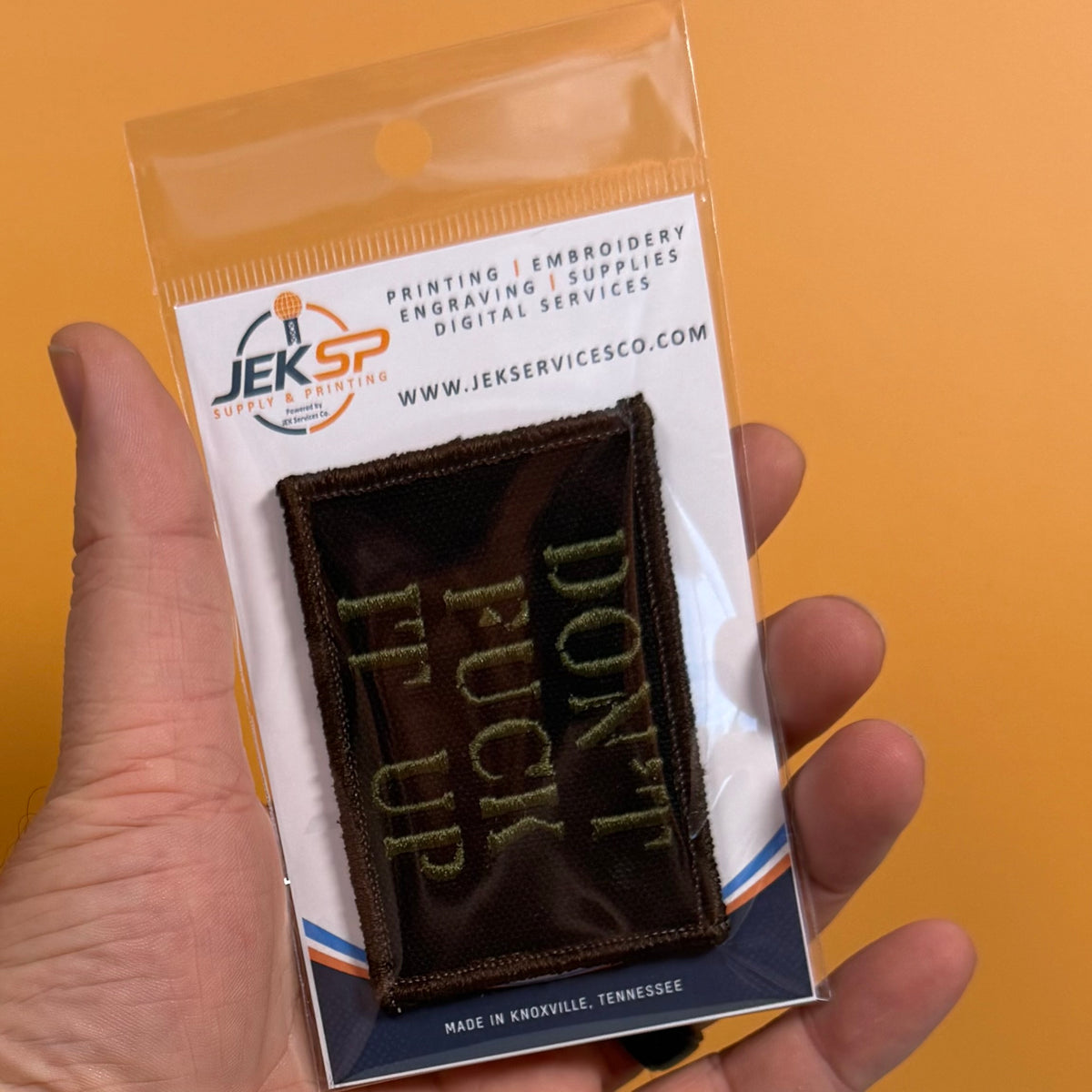 A hand holds a small embroidered patch in clear plastic packaging against a solid orange background. The patch is dark brown with light green text reading 'DON'T FUCK UP.