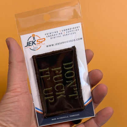A hand holds a small embroidered patch in clear plastic packaging against a solid orange background. The patch is dark brown with light green text reading 'DON'T FUCK UP.
