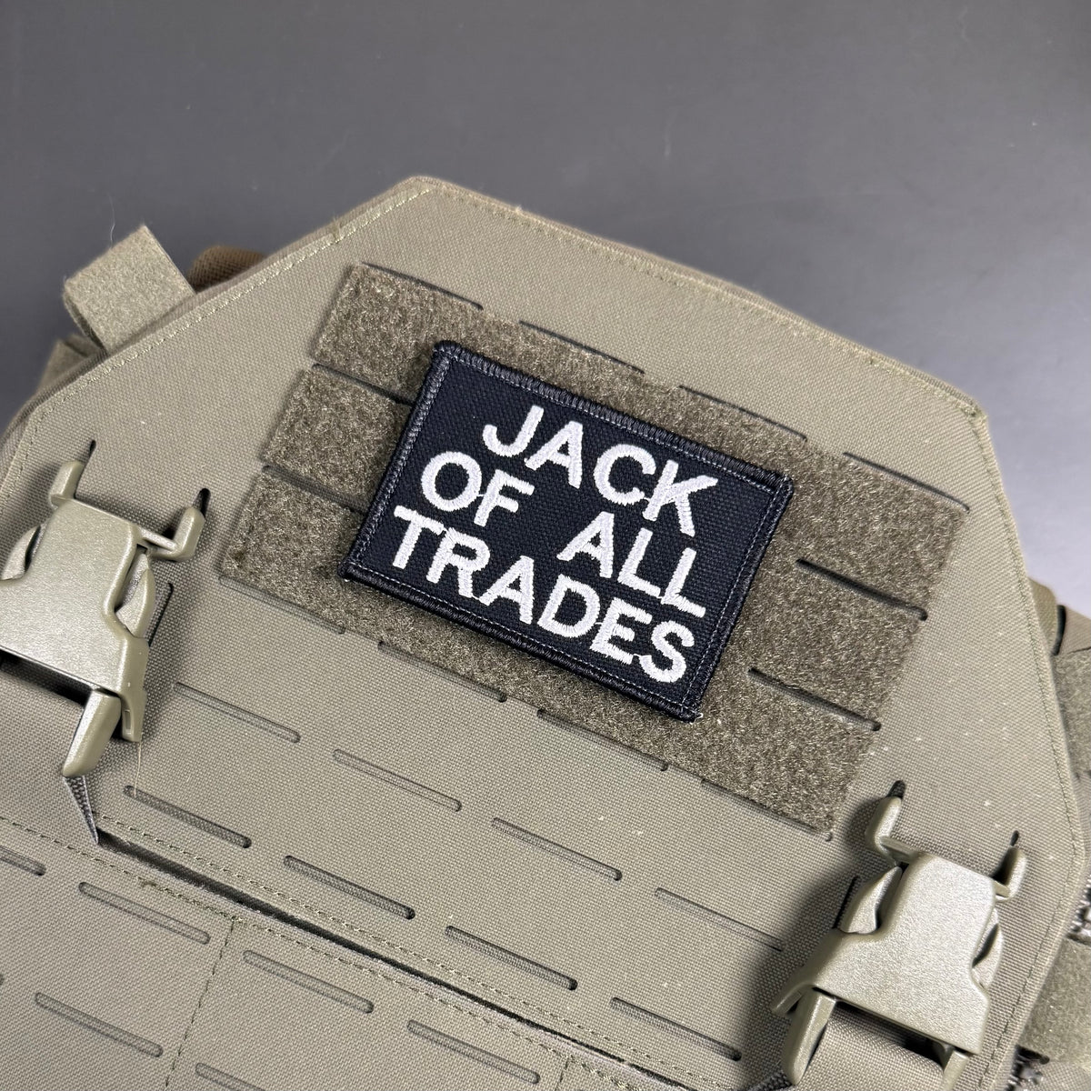 Jack Of All Trades embroidered morale patch: tactical vest or plate carrier in coyote tan with a black rectangular patch reading 'JACK OF ALL TRADES' in white text, attached to hook-and-loop panels.