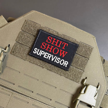 A close-up of a tactical vest or plate carrier in coyote tan, featuring a black rectangular patch with the words 'SHIT SHOW SUPERVISOR' in red and white embroidered lettering, mounted on a hook-and-loop section.