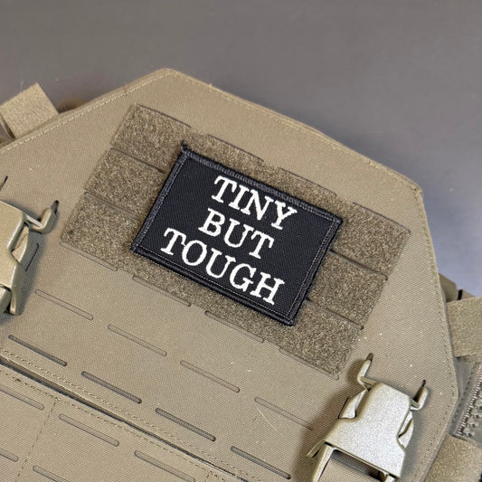 Tiny But Tough embroidered morale patch: tactical vest or plate carrier in coyote tan, featuring a black rectangular embroidered patch with the white text 'TINY BUT TOUGH' on the front hook-and-loop panel.