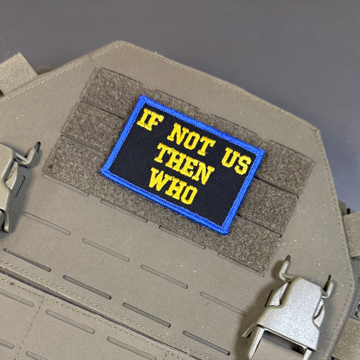 If Not Us Then Who embroidered morale patch: embroidered tactical patch with the text 'IF NOT US THEN WHO' in yellow on a black background with a blue border, shown attached to tan webbing on a tactical vest or plate carrier.