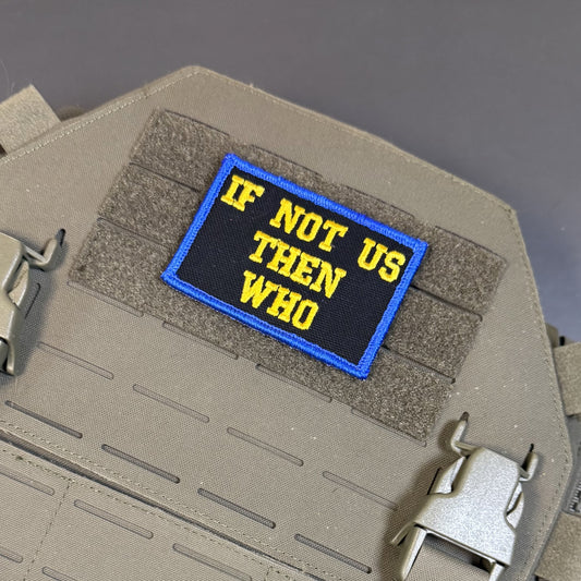 A rectangular embroidered tactical patch with the text 'IF NOT US THEN WHO' in yellow on a black background with a blue border, shown attached to tan webbing on a tactical vest or plate carrier.