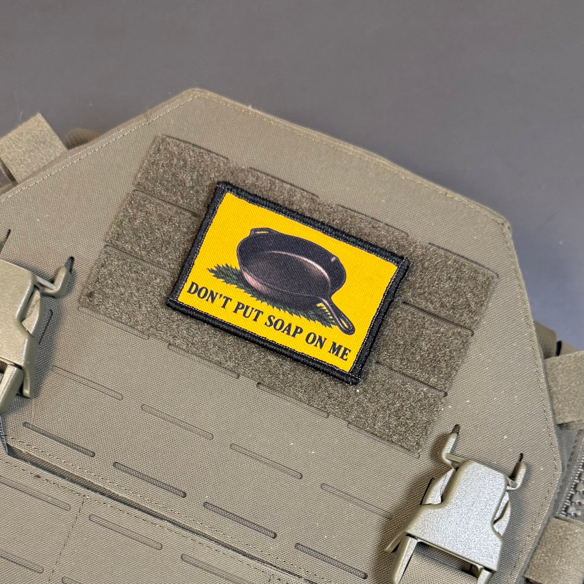 A rectangular tactical patch with a bright yellow background and a black skillet illustration, reading 'DON'T PUT SOAP ON ME,' attached to tan webbing on a tactical vest or plate carrier.