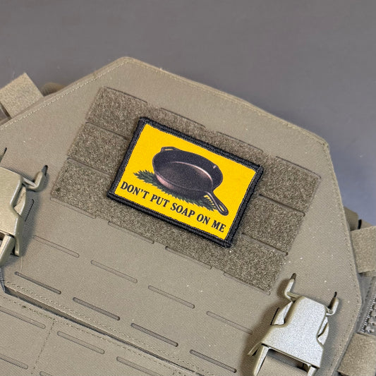 A rectangular tactical patch with a bright yellow background and a black skillet illustration, reading 'DON'T PUT SOAP ON ME,' attached to tan webbing on a tactical vest or plate carrier.