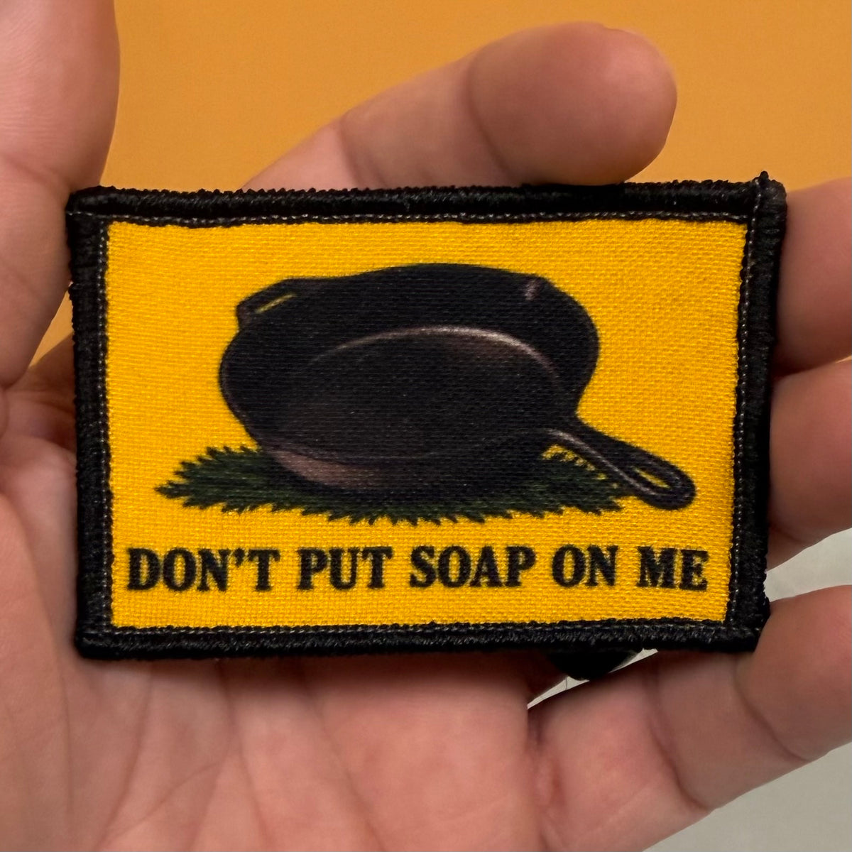 A rectangular embroidered patch held in a hand, featuring a yellow background with a black cast iron skillet resting on green grass and the text 'DON'T PUT SOAP ON ME' in black. The patch has a black stitched border.