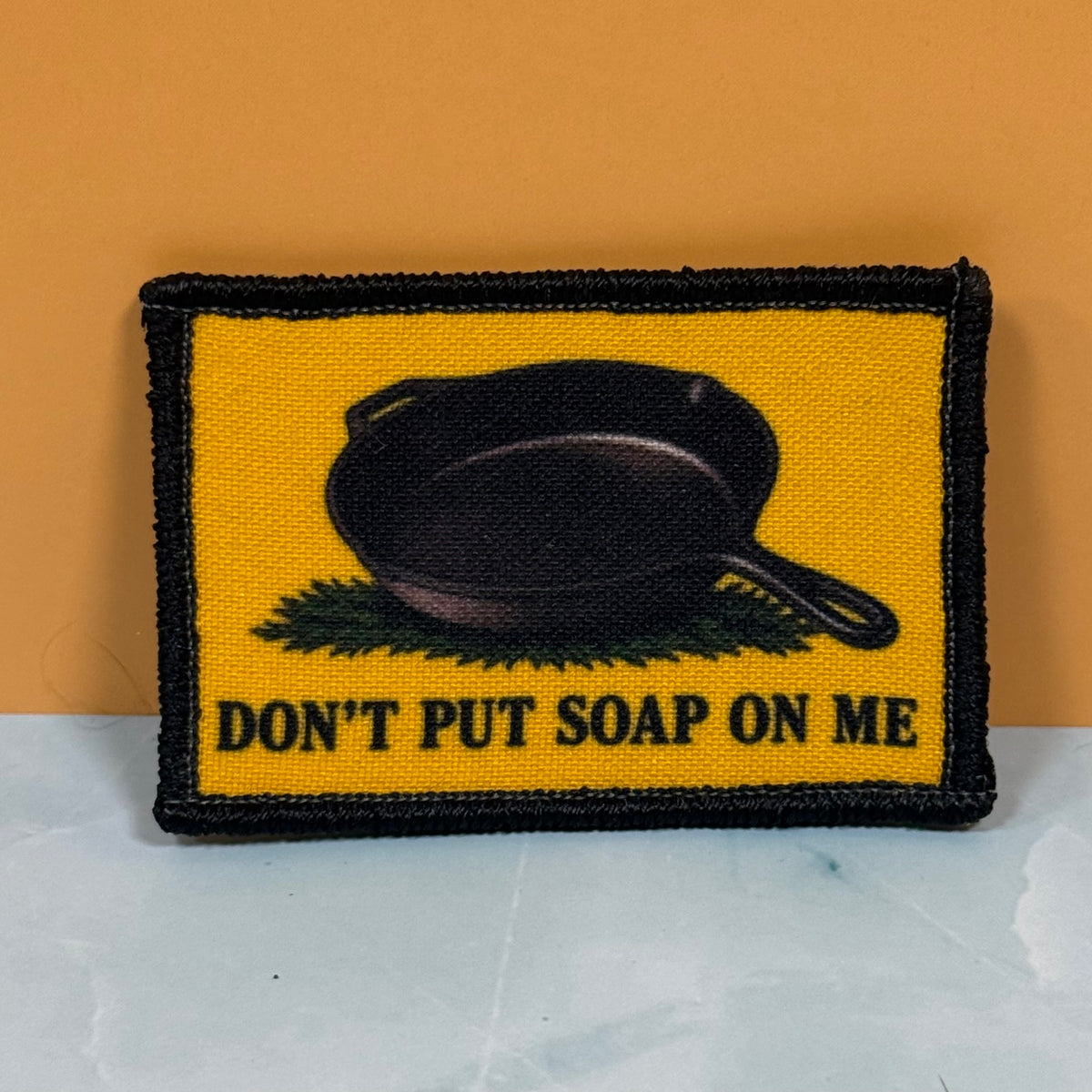 A rectangular embroidered patch with a yellow background, black border, a black cast-iron skillet graphic, and the text 'DON'T PUT SOAP ON ME' in black. The patch is photographed standing upright against an orange wall on a light marble-like surface.