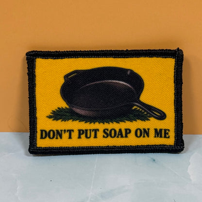 A rectangular embroidered patch with a yellow background, black border, a black cast-iron skillet graphic, and the text 'DON'T PUT SOAP ON ME' in black. The patch is photographed standing upright against an orange wall on a light marble-like surface.