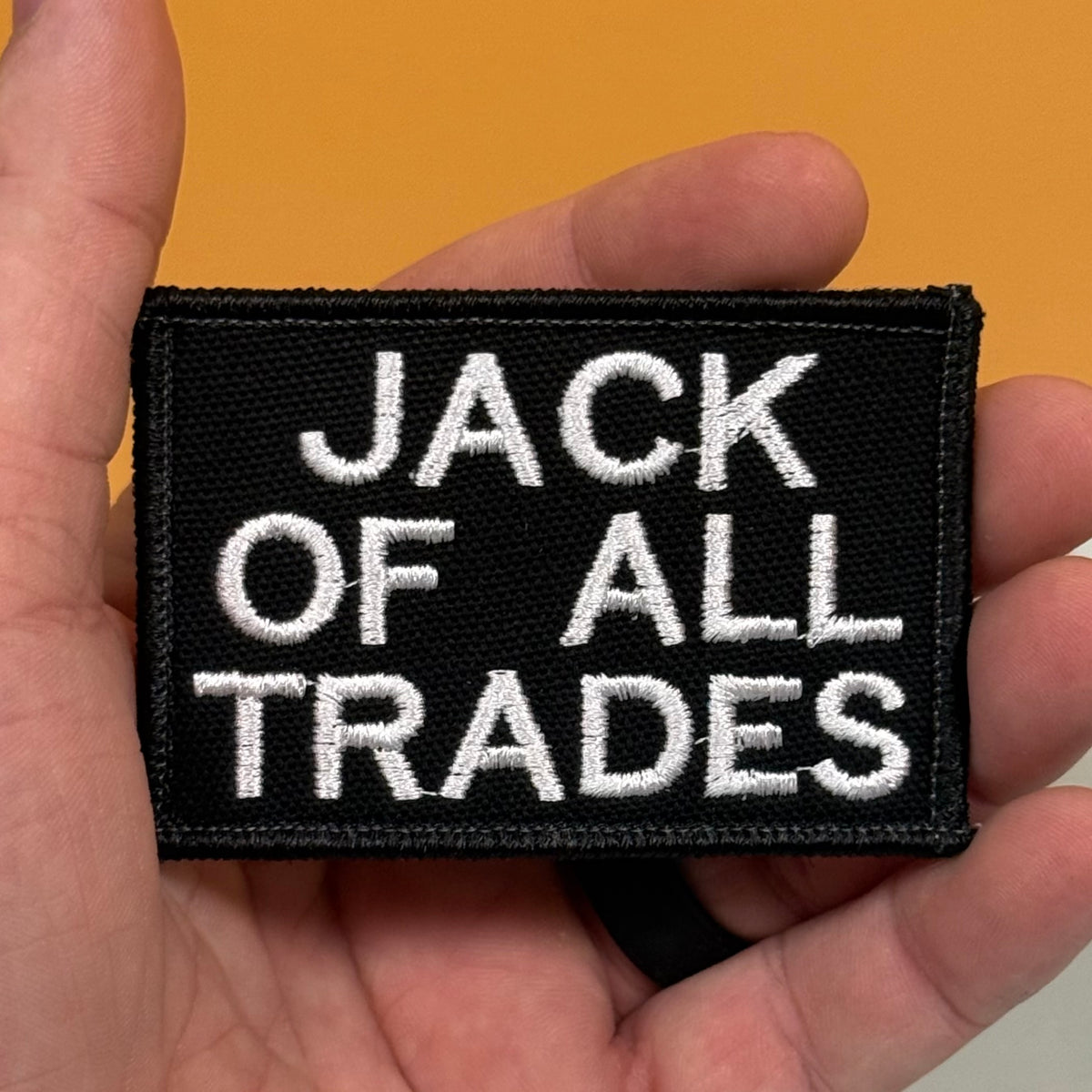 Jack Of All Trades embroidered morale patch: black embroidered patch with white text reading 'JACK OF ALL TRADES' held in a person's hand against a solid orange background.