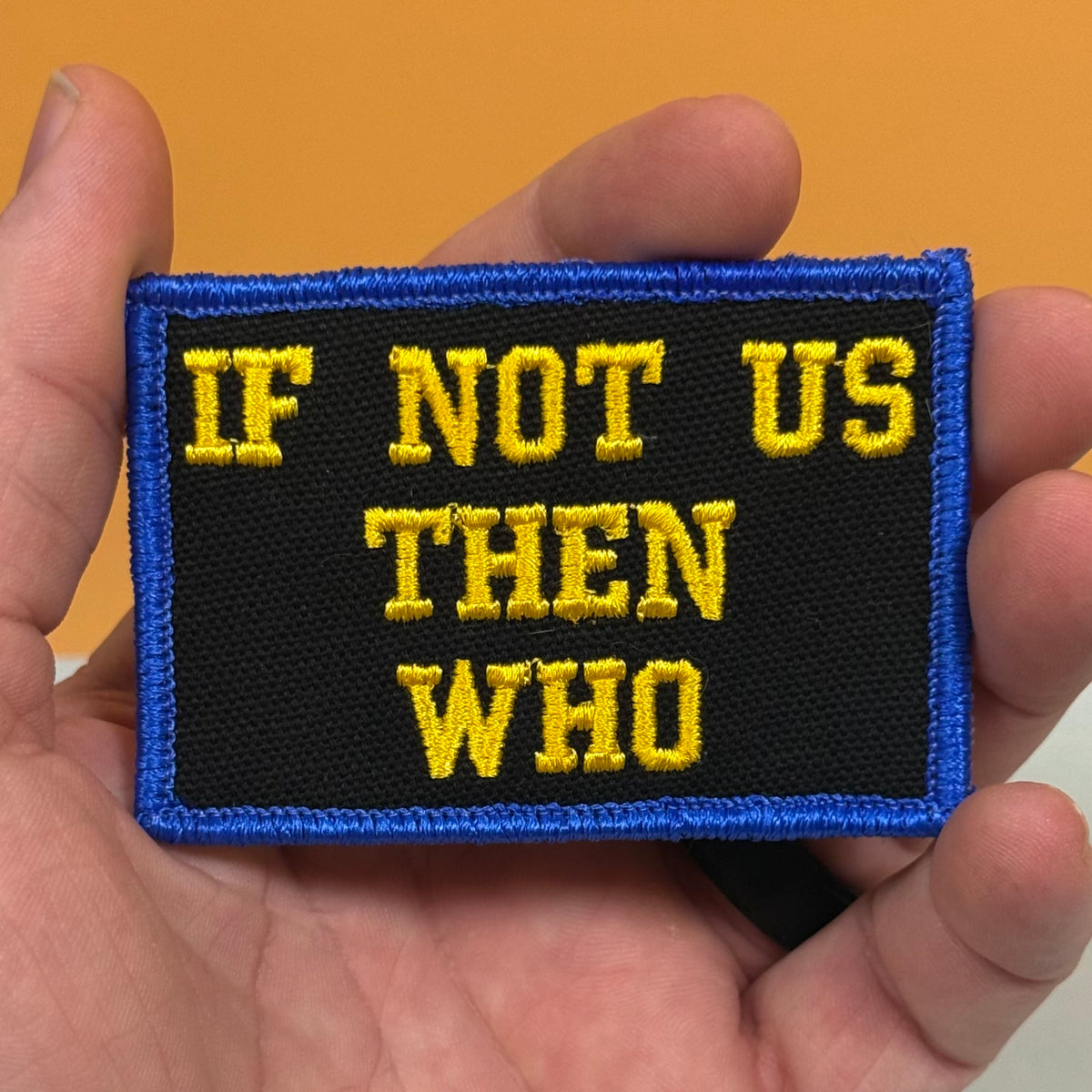 If Not Us Then Who embroidered morale patch: held in a hand, featuring the text 'IF NOT US THEN WHO' in yellow embroidery on a black background with a blue border.