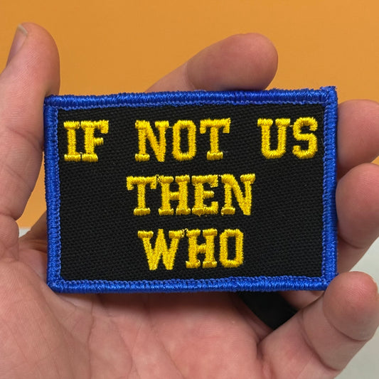 A rectangular embroidered patch held in a hand, featuring the text 'IF NOT US THEN WHO' in yellow embroidery on a black background with a blue border.