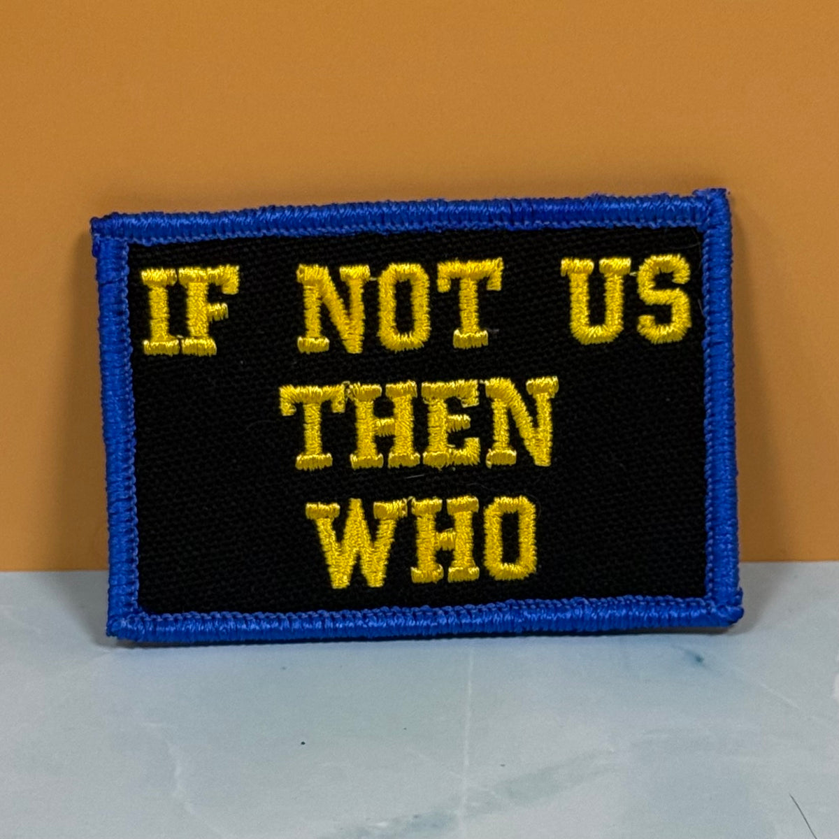 If Not Us Then Who embroidered morale patch: with a black center, blue border, and yellow text reading 'IF NOT US THEN WHO', photographed against a warm orange wall and light gray surface.