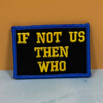 If Not Us Then Who embroidered morale patch: with a black center, blue border, and yellow text reading 'IF NOT US THEN WHO', photographed against a warm orange wall and light gray surface.