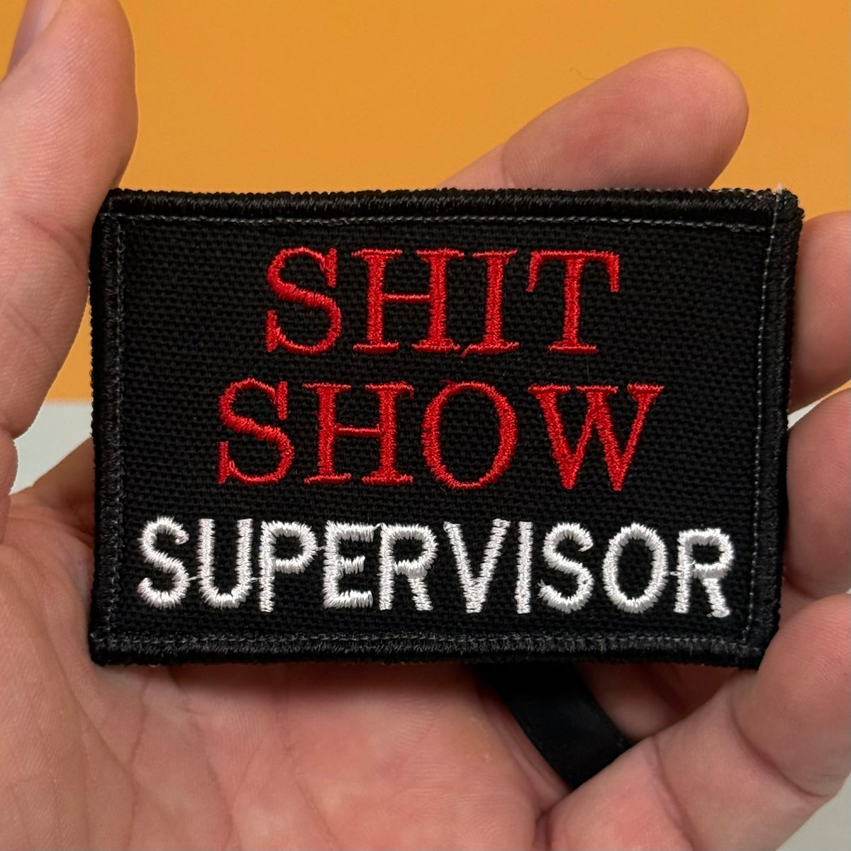 A rectangular embroidered patch held in a hand, featuring the text 'SHIT SHOW SUPERVISOR' in red and white embroidery on a black background.