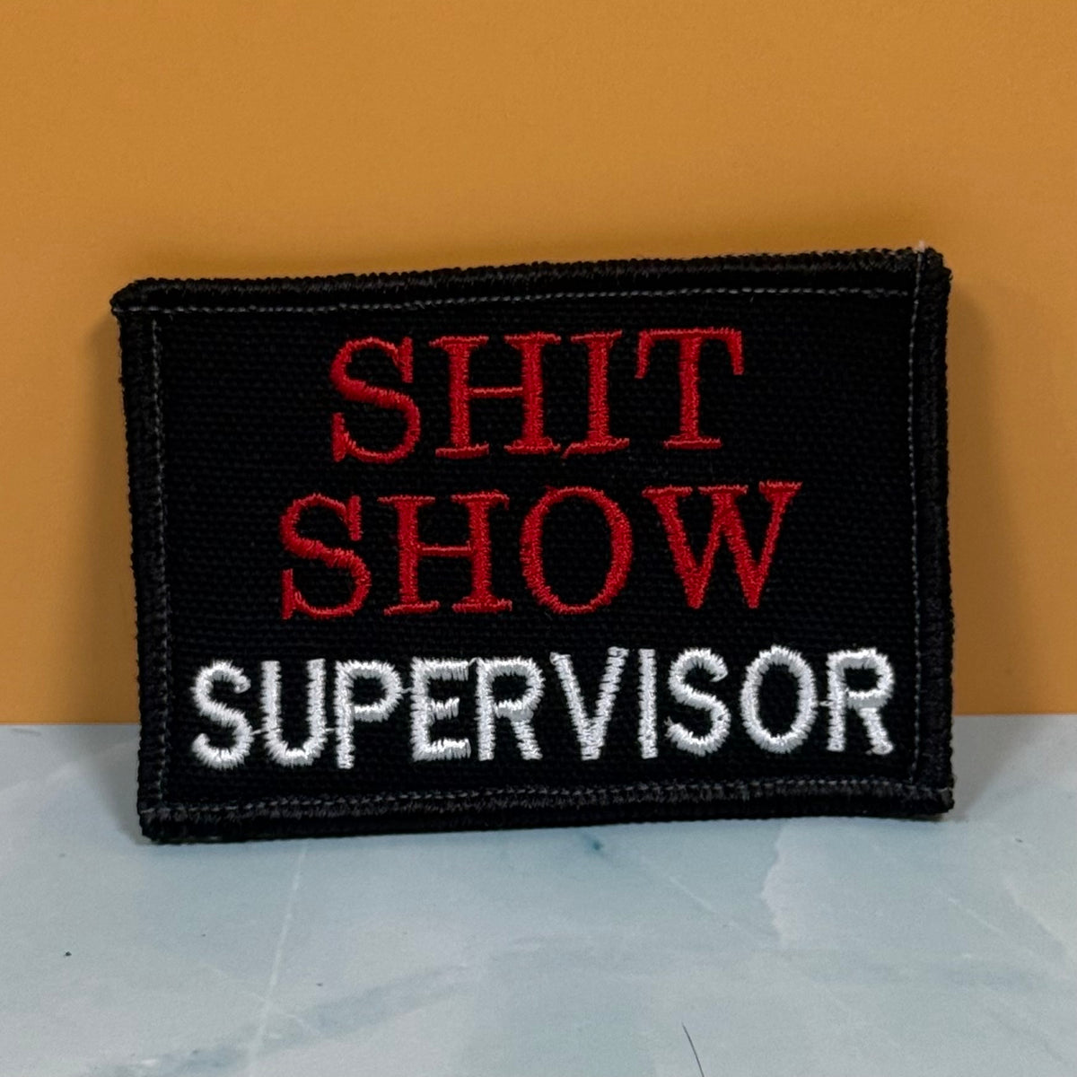 A rectangular black embroidered patch with the words 'SHIT SHOW' in red and 'SUPERVISOR' in white, placed against a mustard yellow background.