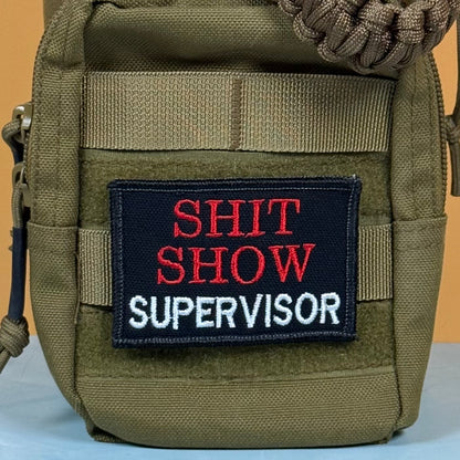 An olive green tactical pouch featuring a black rectangular patch with the embroidered text 'SHIT SHOW SUPERVISOR' in red and white, shown against a solid tan background.